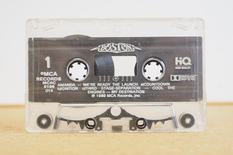 May include: A clear plastic cassette tape case with a black cassette tape inside. The cassette tape has the text "BOSTON" in a silver font on the top. The cassette tape has the text "MCA RECORDS MCAC 6188 014" on the left side and the text "AMANDA - WE'RE READY THE LAUNCH: a) COUNTDOWN IGNITION c) THIRD STAGE SEPARATION - COOL THE ENGINES - MY DESTINATION &copy; 1986 MCA Records, Inc" on the right side. The cassette tape also has the text "HQ HIGH QUALITY" on the right side.
