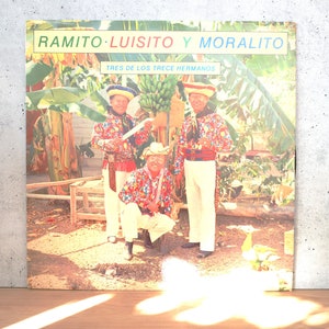 May include: A vintage vinyl record album cover featuring three men in colorful patterned shirts and hats, standing in a banana plantation. The album title is "Ramito Luisito y Moralito" and the subtitle is "Tres de los Trece Hermanos".