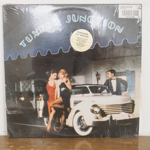 May include: A vinyl record album cover featuring a white car with three people standing next to it. The album cover is for the band "Tuxedo Junction" and the album title is "Contains The Singles".