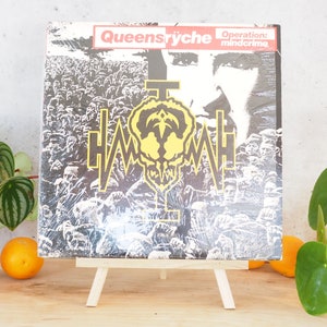 May include: A vinyl record on a wooden easel. The record cover is black and white with a yellow and black logo. The album title is "Operation: Mindcrime" by Queensrÿche.