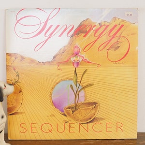 May include: A vintage vinyl record album cover featuring a desert landscape with a mountain range in the background. The album title "Synergy" is written in pink cursive lettering at the top of the cover. The album title "Sequencer" is written in red lettering at the bottom of the cover. The album is by the artist "Laszlo".