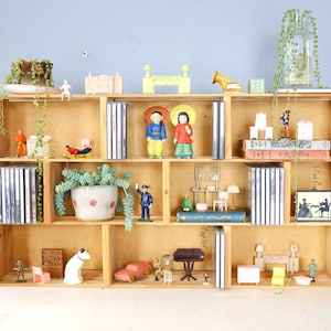 May include: A wooden display shelf with multiple compartments, filled with various decorative items. Includes miniature figurines, potted plants, books, CDs, and small furniture pieces. The shelf is against a blue wall.