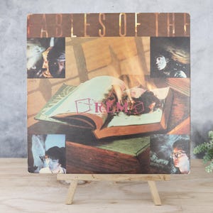 May include: Album cover for "Fables of the Reconstruction" by R.E.M. features a collage of images, including portraits and an open book with flames. The album is displayed on a small wooden easel against a gray background.