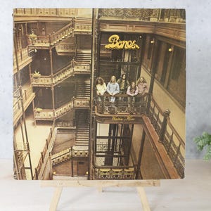 May include: Album cover featuring a group of people on a balcony inside a building with multiple staircases. The album cover has the word "Bones" in a gold color. The album is displayed on a wooden easel.