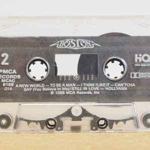 May include: A clear plastic cassette tape case with a black cassette tape inside. The cassette tape has the band name "BOSTON" printed on it in white letters. The tape is labeled with the song titles "A New World", "To Be A Man", "I Think I Like It", "Can'tcha Say", "Still In Love", and "Hollyann". The tape is from MCA Records and is labeled as "High Quality".