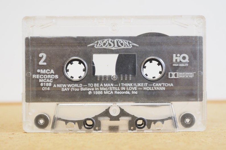 May include: A clear plastic cassette tape case with a black cassette tape inside. The cassette tape has the band name "BOSTON" printed on it in white letters. The tape is labeled with the song titles "A New World", "To Be A Man", "I Think I Like It", "Can'tcha Say", "Still In Love", and "Hollyann". The tape is from MCA Records and is labeled as "High Quality".