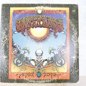 May include: Vintage vinyl record album cover featuring psychedelic artwork. The cover displays a skull with crossed bones, a sunburst, and ornate lettering. The color palette includes orange, blue, and brown tones.