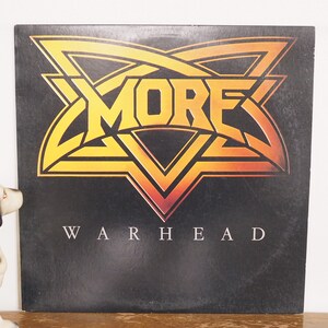 May include: A black vinyl record with a yellow and orange starburst design. The band name "MORE" is written in a stylized font inside the starburst. The album title "WARHEAD" is written in white letters below the starburst.