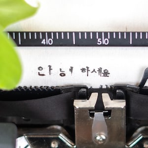 Könnte beinhalten: Nahaufnahme einer alten Schreibmaschine mit dem koreanischen Satz "안녕하세요" auf einem weißen Blatt Papier. Die Schreibmaschine ist schwarz und silberfarben, mit einer linealartigen Skala über dem Text. Ein grünes Blatt befindet sich in der oberen linken Ecke.
