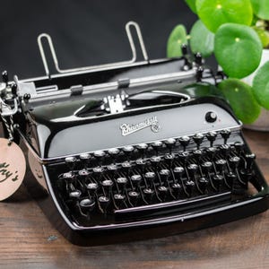 Könnte beinhalten: Eine glänzend schwarze Rheinmetall-Schreibmaschine mit QWERTY-Tastaturlayout und einem runden Holzanhänger an der Seite, auf dem der Name Kathy's in schwarzer Schreibschrift steht. Das Gerät verfügt über silberne Metallakzente und einen Papierhalter.