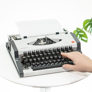 May include: A classic Olympia typewriter in light grey with black keys, and a sheet of white paper. A hand is poised above the keys. The word "Olympia" is printed on the front of the machine. The typewriter is on a white table.