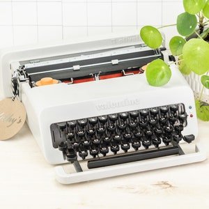 Op de afbeelding: Een witte vintage typemachine met een zwart toetsenbord en een bruin papieren label met de tekst "Kathy's".