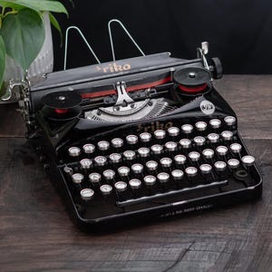May include: A vintage black Erika typewriter with white keys and red ribbon spools. The typewriter has the brand name "Erika" on the top and is set on a dark wooden surface. A potted plant with green leaves is in the background.