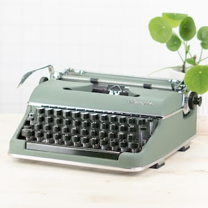 Könnte beinhalten: Eine vintage Schreibmaschine in Salbeigrün mit schwarzen Tasten und silbernen Akzenten. Die Schreibmaschine steht auf einer hellen Holzoberfläche, im Hintergrund eine kleine Topfpflanze. Die Tasten sind in einem Standard-QWERTZ-Layout angeordnet.