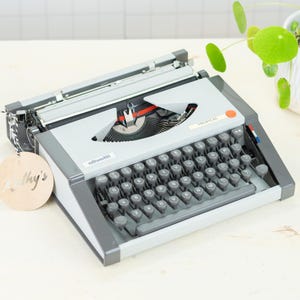 Peut inclure: Une machine à écrire Olivetti vintage grise et noire, avec un clavier complet et un mécanisme d'alimentation du papier. La machine à écrire a une étiquette ronde en bois qui dit "Kathy's". Une plante verte dans un pot blanc est en arrière-plan.