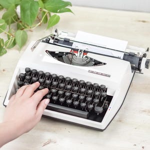 Puede incluir: Una máquina de escribir manual blanca con teclas circulares negras y distribución QWERTY. La máquina cuenta con un carro de metal, un alimentador de papel con una hoja blanca insertada y el nombre de la marca Pippa impreso en plata en la carcasa.