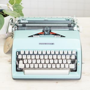 Op de afbeelding: Een vintage lichtblauwe Olympia-Colortip-S schrijfmachine. De schrijfmachine heeft witte toetsen en een chromen wagen. De afbeelding toont de voorkant van de schrijfmachine, met het toetsenbord en het Olympia-Colortip-S logo.