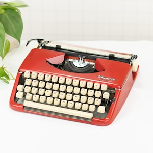 Peut inclure: Une machine à écrire Olympia rouge vintage avec des touches blanc cassé. La machine à écrire présente un design classique avec un levier de retour de chariot et un rouleau d'alimentation du papier. Les touches sont disposées en QWERTY. La marque "Olympia" est visible.