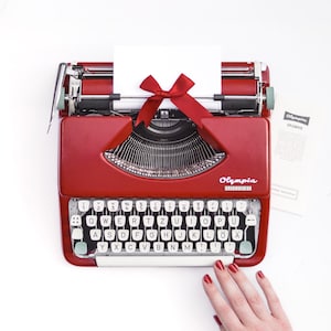 Könnte beinhalten: Eine rote Olympia Splendid 33 Schreibmaschine mit eingelegtem weißem Blatt Papier, verziert mit einer dunkelroten Satinschleife. Die Schreibmaschine verfügt über ein QWERTZ-Tastenlayout mit weißen Tasten und einen silbernen Wagenrücklaufhebel.