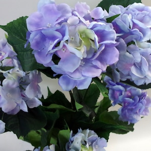 May include: A bouquet of artificial hydrangea flowers in shades of light blue and purple. The flowers are arranged in a cluster with green leaves.