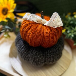 May include: Two knitted pumpkins, one orange and one gray, with a white lace bow on the orange pumpkin. The pumpkins are sitting on a wooden tray.