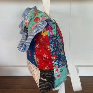 Puede incluir: Una mochila de patchwork con una variedad de telas estampadas florales y de otros tipos en rojo, azul y verde azulado. La bolsa tiene un ribete de cuadros azules y blancos y una correa blanca. Una cremallera es visible en el lateral.