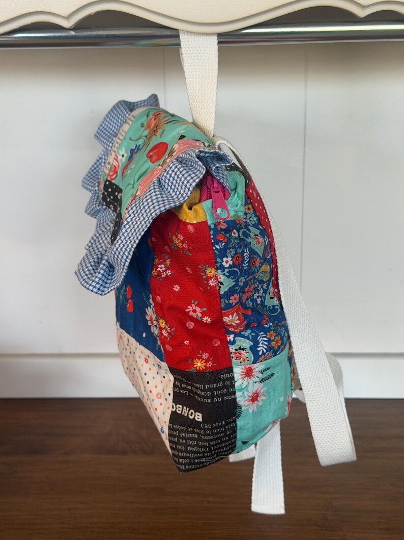 Puede incluir: Una mochila de patchwork con una variedad de telas estampadas florales y de otros tipos en rojo, azul y verde azulado. La bolsa tiene un ribete de cuadros azules y blancos y una correa blanca. Una cremallera es visible en el lateral.