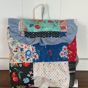 May include: A patchwork backpack with a variety of fabric patterns, including cherries, flowers, and polka dots. The top features a ruffled edge and a white handle. The bag has white straps.