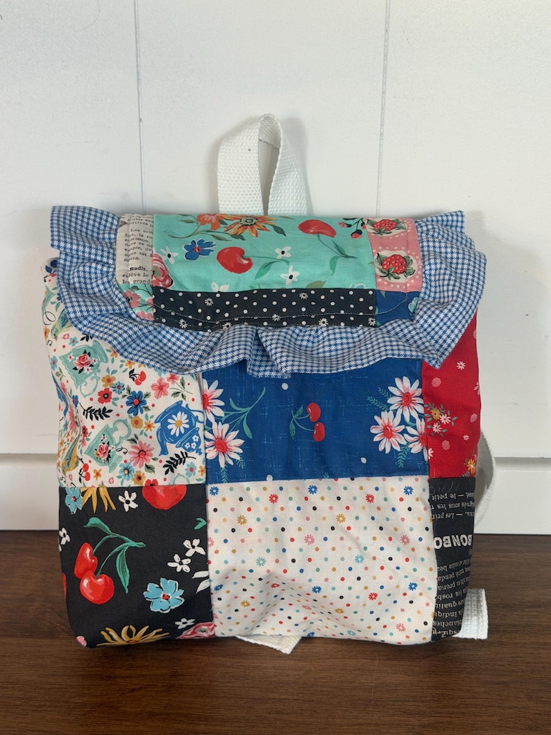 Puede incluir: Una mochila de patchwork con una variedad de patrones de tela, incluyendo cerezas, flores y lunares. La parte superior presenta un borde con volantes y un asa blanca. La bolsa tiene correas blancas.