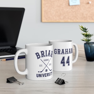 Puede incluir: Dos tazas de cerámica blanca. Una taza presenta el texto "Brian Graham Hawks University" con palos de hockey cruzados. La otra taza tiene el nombre "Graham" y el número "44" en azul marino.