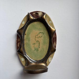 Puede incluir: Un aplique de pared ovalado de cerámica con un panel central de color verde claro agrietado y una pequeña ilustración de una nube verde. La pieza está enmarcada por un borde de bronce metálico oscuro con puntas y una pequeña repisa.