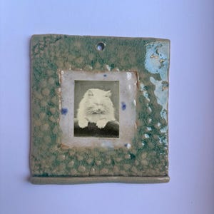 May include: A square ceramic wall hanging with a textured, crackled green and beige glaze. The centre features a rectangular inset containing a black and white photograph of a fluffy cat with its eyes closed. A small hole is at the top centre.