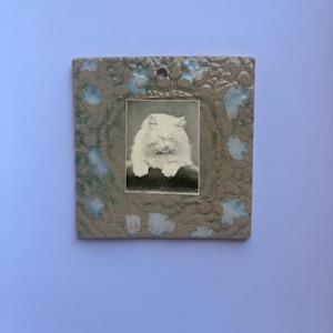 May include: A square ceramic wall plaque featuring a central black and white photograph of a fluffy cat with closed eyes. The plaque has a textured, mottled beige and light blue glaze finish and includes a small circular hole at the top center.
