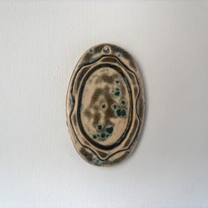 Puede incluir: Adorno de pared ovalado de cerámica con una superficie texturizada y moteada. La pieza presenta un borde marrón oscuro y un centro más claro, de color beige, con detalles en verde oscuro. Se ve un pequeño agujero en la parte superior para colgar.