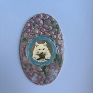 Puede incluir: Un adorno de cerámica de forma ovalada con una superficie texturizada y rugosa, con un esmalte moteado en tonos violeta, rosa y verde. Una inserción circular en el centro muestra una fotografía vintage en blanco y negro de un gato blanco.