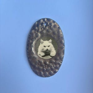 May include: An oval-shaped pendant with a hammered, metallic bronze-toned finish featuring a circular inset image of a fluffy white cat. The pendant has a small hole at the top for hanging and displays a textured, uneven surface throughout the frame.