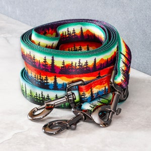 Op de afbeelding: Een kleurrijke hondenriem met een bosprint en twee zilveren metalen clips. De riem is gemaakt van duurzaam nylon en heeft een levendig ontwerp met bomen, bergen en een zonsondergang.