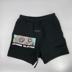 May include: Black shorts with a drawstring waist and the word "ESSENTIALS" printed on the right leg. A rectangular patch on the left leg features a cartoon illustration of a character from "Demon Slayer" with the text "DEMON SLAYER".