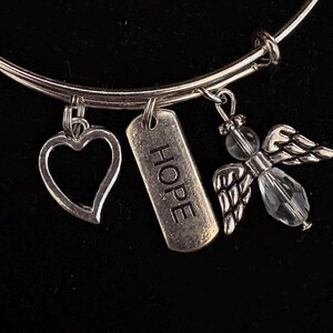 Puede incluir: Pulsera de brazalete plateada con tres colgantes. Los colgantes incluyen un coraz&oacute;n, una etiqueta rectangular con la palabra "HOPE" y un &aacute;ngel con una cuenta de cristal y alas detalladas. Sobre un fondo negro.