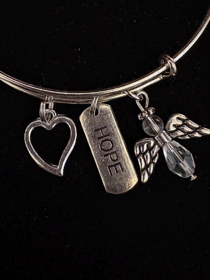 Puede incluir: Pulsera de brazalete plateada con tres colgantes. Los colgantes incluyen un coraz&oacute;n, una etiqueta rectangular con la palabra "HOPE" y un &aacute;ngel con una cuenta de cristal y alas detalladas. Sobre un fondo negro.