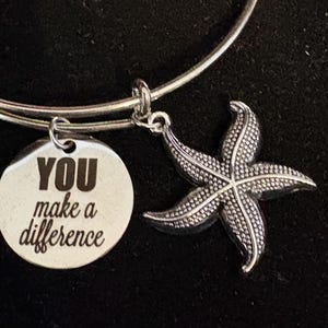Puede incluir: Pulsera de brazalete plateada con un dije de estrella de mar y un dije redondo con el texto "YOU make a difference". El dije de estrella de mar tiene una superficie texturizada. La pulsera se muestra sobre un fondo oscuro.