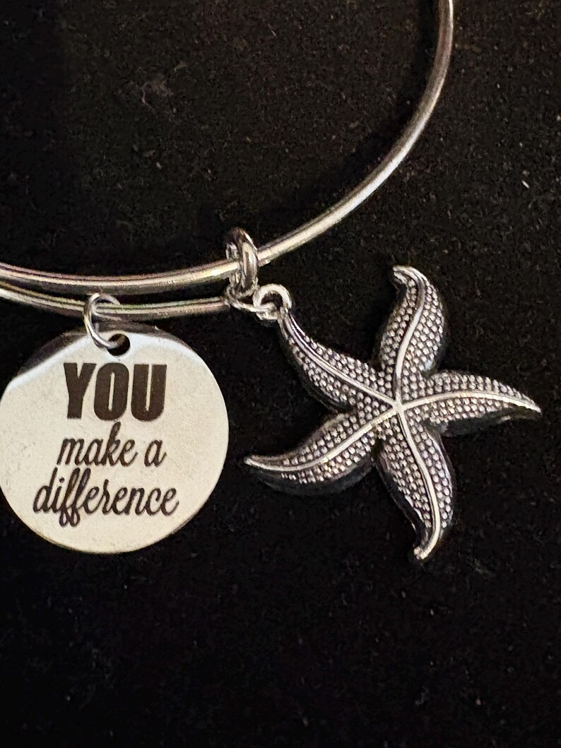 Puede incluir: Pulsera de brazalete plateada con un dije de estrella de mar y un dije redondo con el texto "YOU make a difference". El dije de estrella de mar tiene una superficie texturizada. La pulsera se muestra sobre un fondo oscuro.