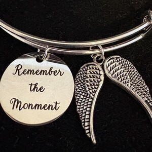 Puede incluir: Pulsera de brazalete plateada con un dije redondo y dos dijes de alas. El dije redondo est&aacute; grabado con las palabras "Remember the Moment". Las alas est&aacute;n detalladas con patrones de plumas. La pulsera se muestra sobre un fondo oscuro.