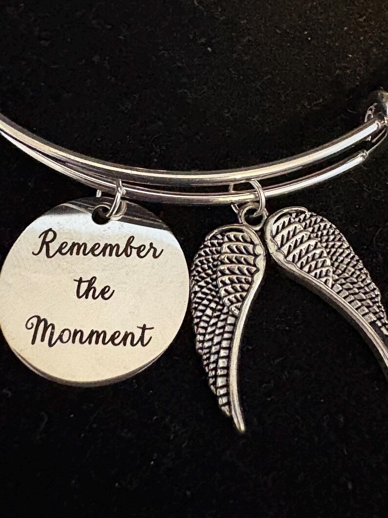Puede incluir: Pulsera de brazalete plateada con un dije redondo y dos dijes de alas. El dije redondo est&aacute; grabado con las palabras "Remember the Moment". Las alas est&aacute;n detalladas con patrones de plumas. La pulsera se muestra sobre un fondo oscuro.