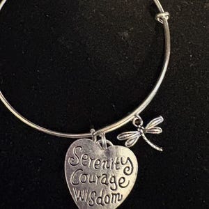 Puede incluir: Pulsera de brazalete plateada con un dije en forma de coraz&oacute;n y un dije de lib&eacute;lula. El dije de coraz&oacute;n tiene grabadas las palabras "Serenity Courage Wisdom". La pulsera est&aacute; sobre un fondo negro.