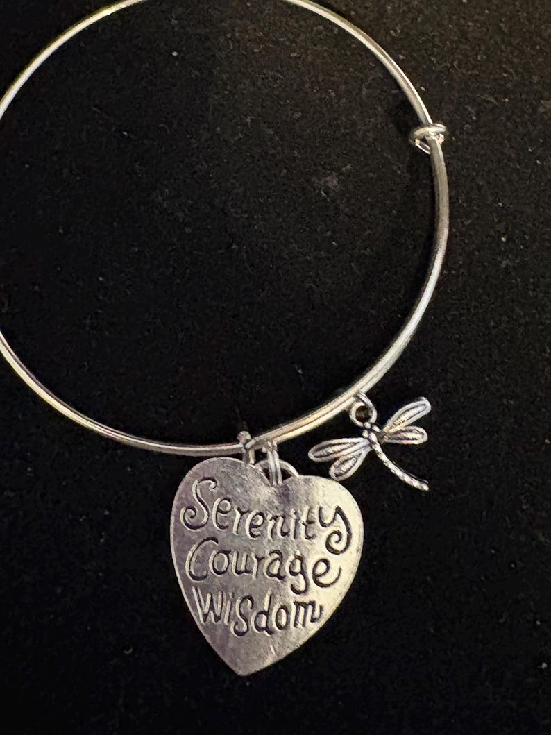 Puede incluir: Pulsera de brazalete plateada con un dije en forma de coraz&oacute;n y un dije de lib&eacute;lula. El dije de coraz&oacute;n tiene grabadas las palabras "Serenity Courage Wisdom". La pulsera est&aacute; sobre un fondo negro.
