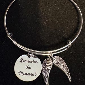 Puede incluir: Pulsera de brazalete plateada con dos colgantes. Un colgante es un disco redondo con las palabras "Remember the Monment" grabadas. El otro colgante es un par de alas de &aacute;ngel detalladas. La pulsera se muestra sobre un fondo negro.