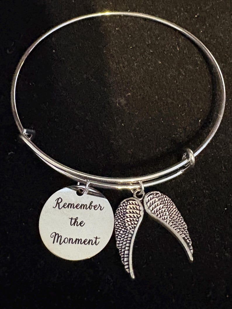 Puede incluir: Pulsera de brazalete plateada con dos colgantes. Un colgante es un disco redondo con las palabras "Remember the Monment" grabadas. El otro colgante es un par de alas de &aacute;ngel detalladas. La pulsera se muestra sobre un fondo negro.