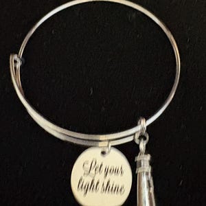 Puede incluir: Pulsera de brazalete plateada con un dije redondo y un dije de faro. El dije redondo tiene grabado el texto "Let your light shine". El dije de faro es detallado y cuelga debajo de la pulsera.