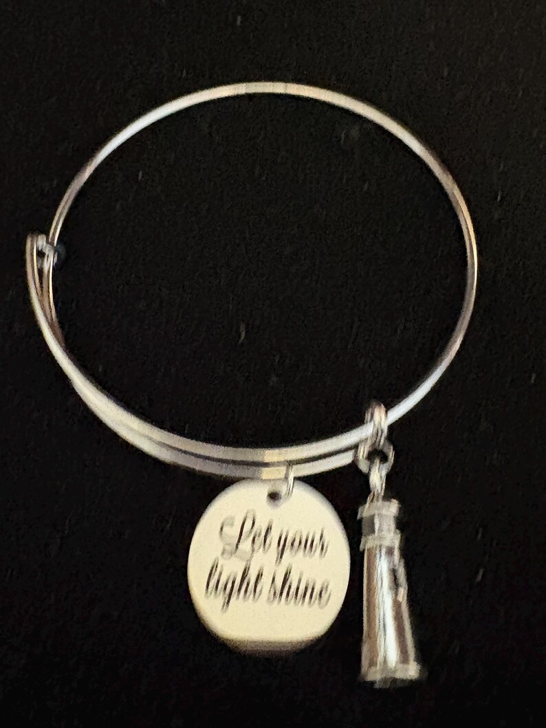 Puede incluir: Pulsera de brazalete plateada con un dije redondo y un dije de faro. El dije redondo tiene grabado el texto "Let your light shine". El dije de faro es detallado y cuelga debajo de la pulsera.